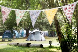 A string of multicoloured, patterned bunting is seen in front of a campsite. Grey and blue tents and wooden logs are seen in the background.