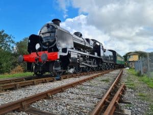 A black steam train with a red bumper plate rolls past the camera, under a blue sky.