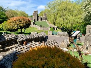 Miniature houses curve down a small grassy hill, next to a miniature model of a church, surrounded by green trees.