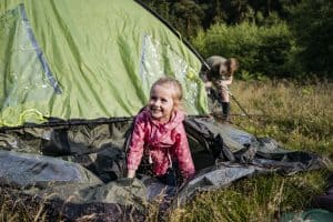 A girl in a pink jacket crawls on a tent ground sheet in front of a green tent