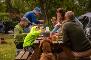 Family enjoy picnic and barbecue food around a brown picnic bench.