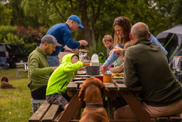 Family enjoy picnic and barbecue food around a brown picnic bench.